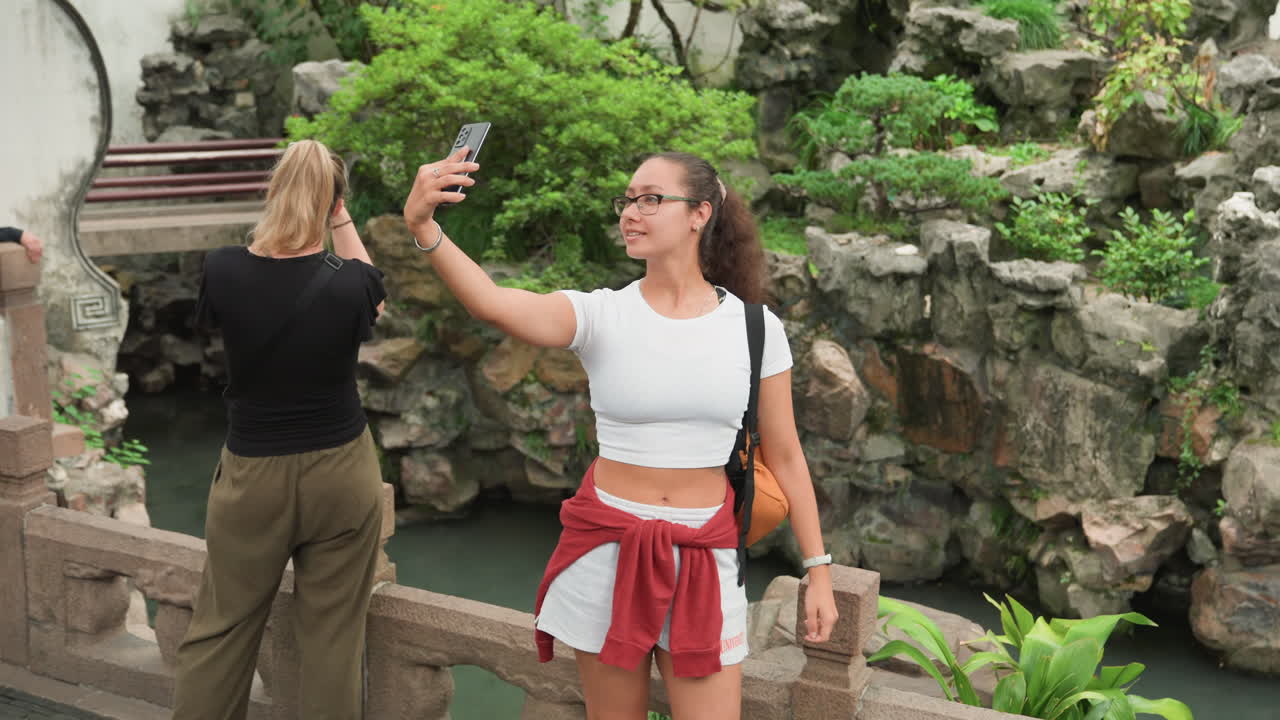 Giovane donna che si scatta un selfie vicino a uno stagno in un giardino roccioso, con un crop top bianco e una giacca rossa legata in vita, con uno zaino, che sorride verso la fotocamera dello smartphone mentre un compagno è di spalle vicino a una ringhiera in pietra.