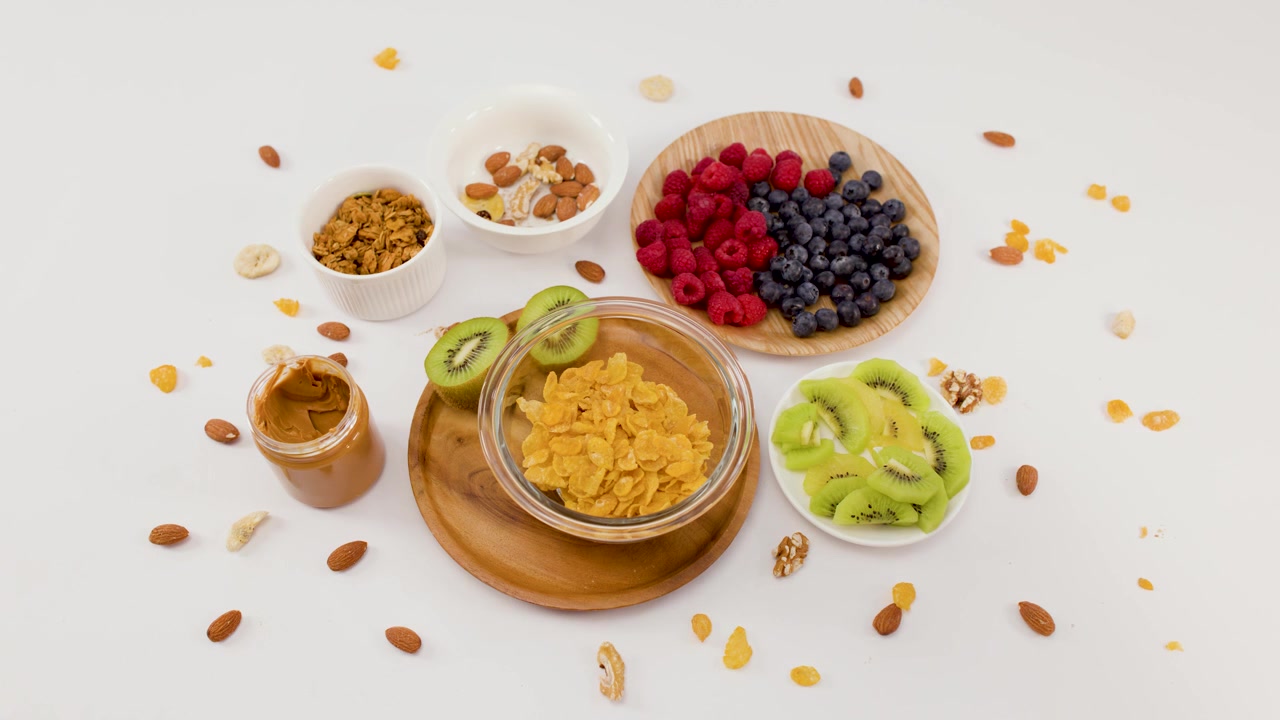Toma aérea de una mano vertiendo cereal en un tazón rodeado de fruta fresca y frutos secos.