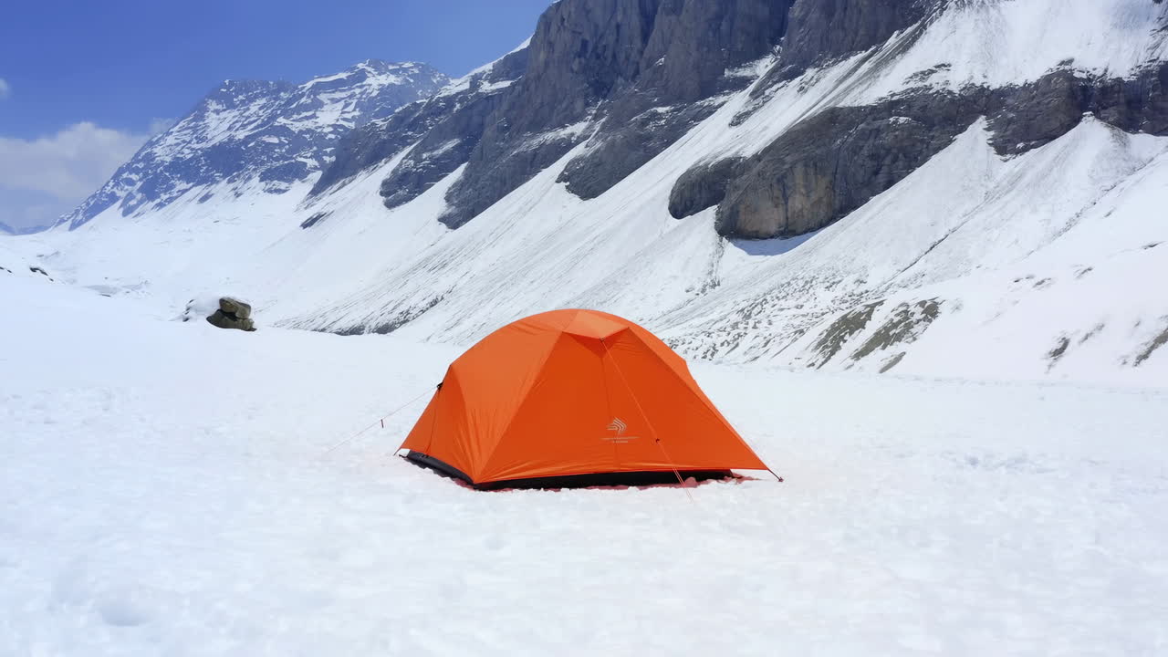 Orange tält på snöig alpin platå, avlägset bergsläger under klarblå himmel, tornande granitklippor.