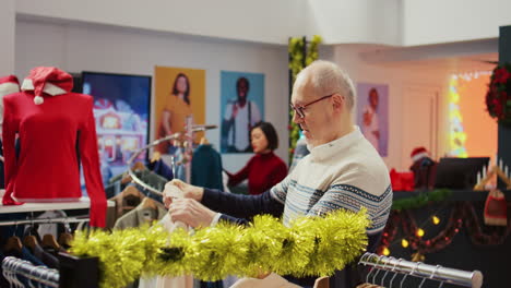 dolly out shot of senescent man in festive decorated clothing store trying to determine if luxurious blazer is the right fit. older client in christmas adorn fashion shop during holiday sales