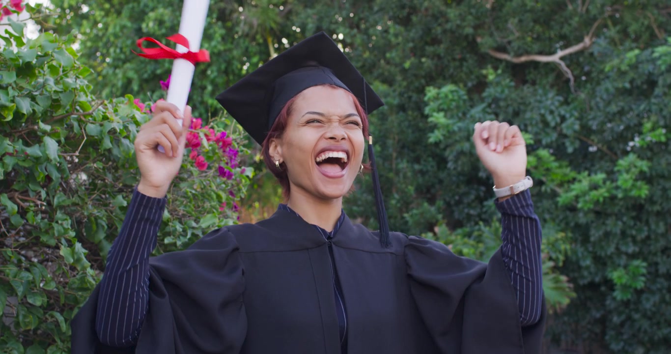 Donna afroamericana che festeggia la laurea, tiene in mano il diploma, saluta e ride, in toga e tocco, in giardino.