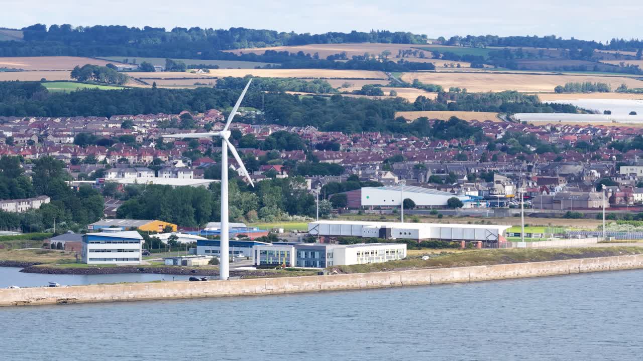 Large wind turbine spins beside water, industrial buildings, and distant fields under daylight