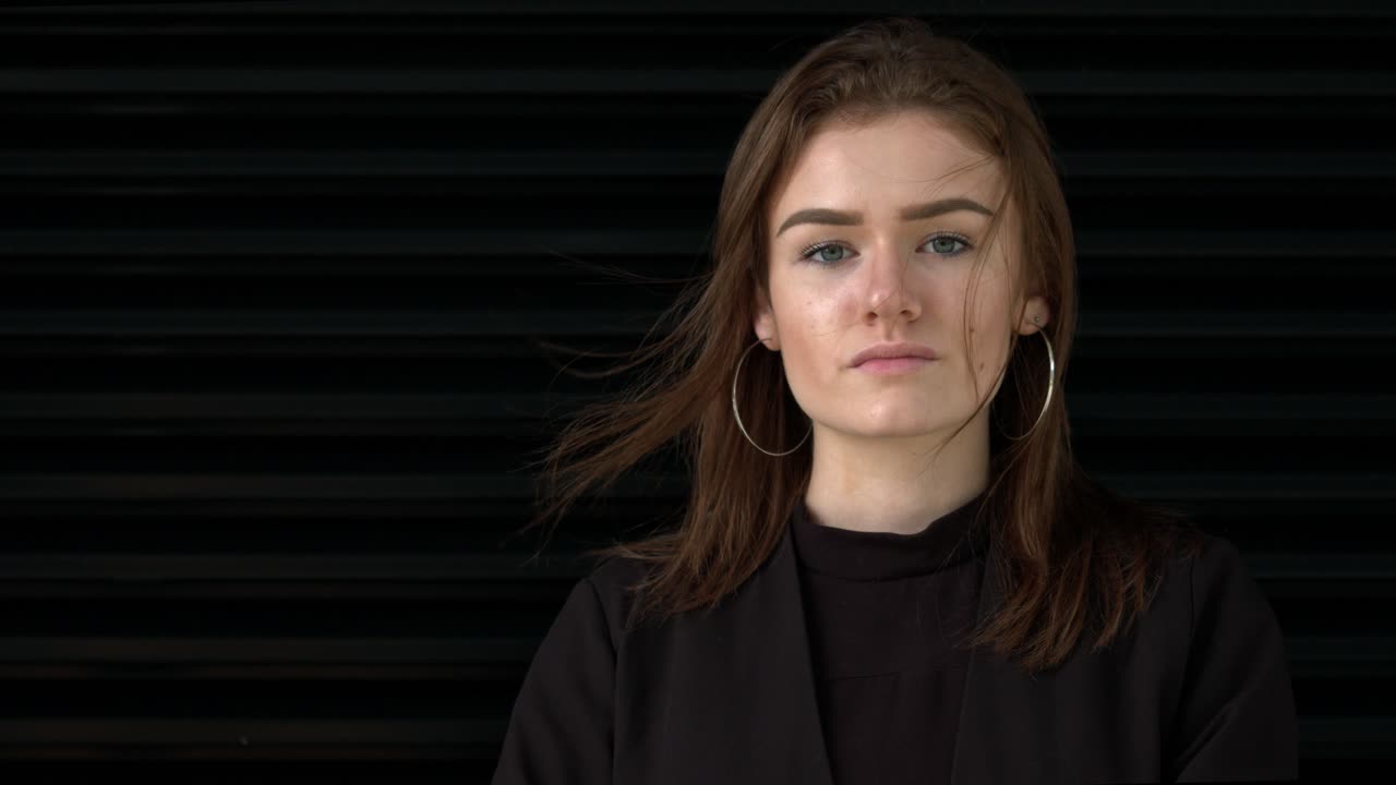 modelo de morena femenina de 20 años mirando seriamente a la cámara, pelo soplando en el viento, fondo negro, cámara lenta