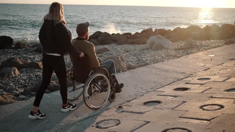 couple supporting a person in a wheelchair on a beach at sunset