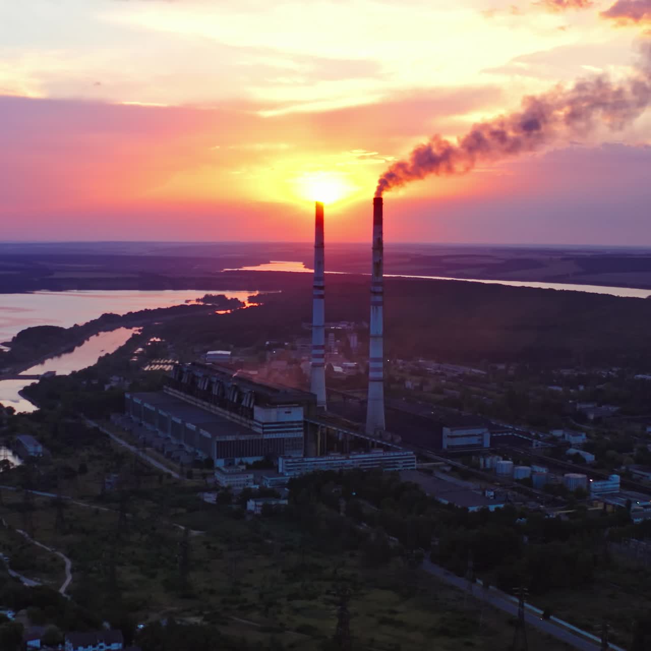 Ecological disaster. Harmful factory near the river releases dirty emissions at sunset. Industry pollute the environment. Pipes with smoke on beautiful nature background.