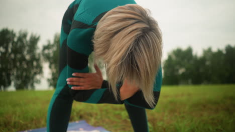 mujer con los brazos plegados giros a la izquierda y a la derecha mientras practica yoga en un vasto campo de hierba, su cabello cubriendo su cara y una alfombra de yoga borrosa debajo de ella, el fondo presenta un campo de herba