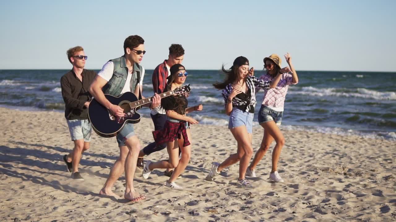 Group of young hipster friends walking and dancing together playing guitar and singing songs on a beach at the water's edge. Slowmotion shot