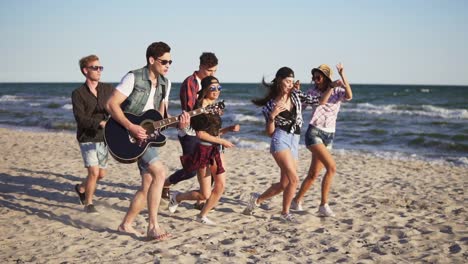 Group-of-young-hipster-friends-walking-and-dancing-together-playing-guitar-and-singing-songs-on-a-beach-at-the-water's-edge.-Slowmotion-shot