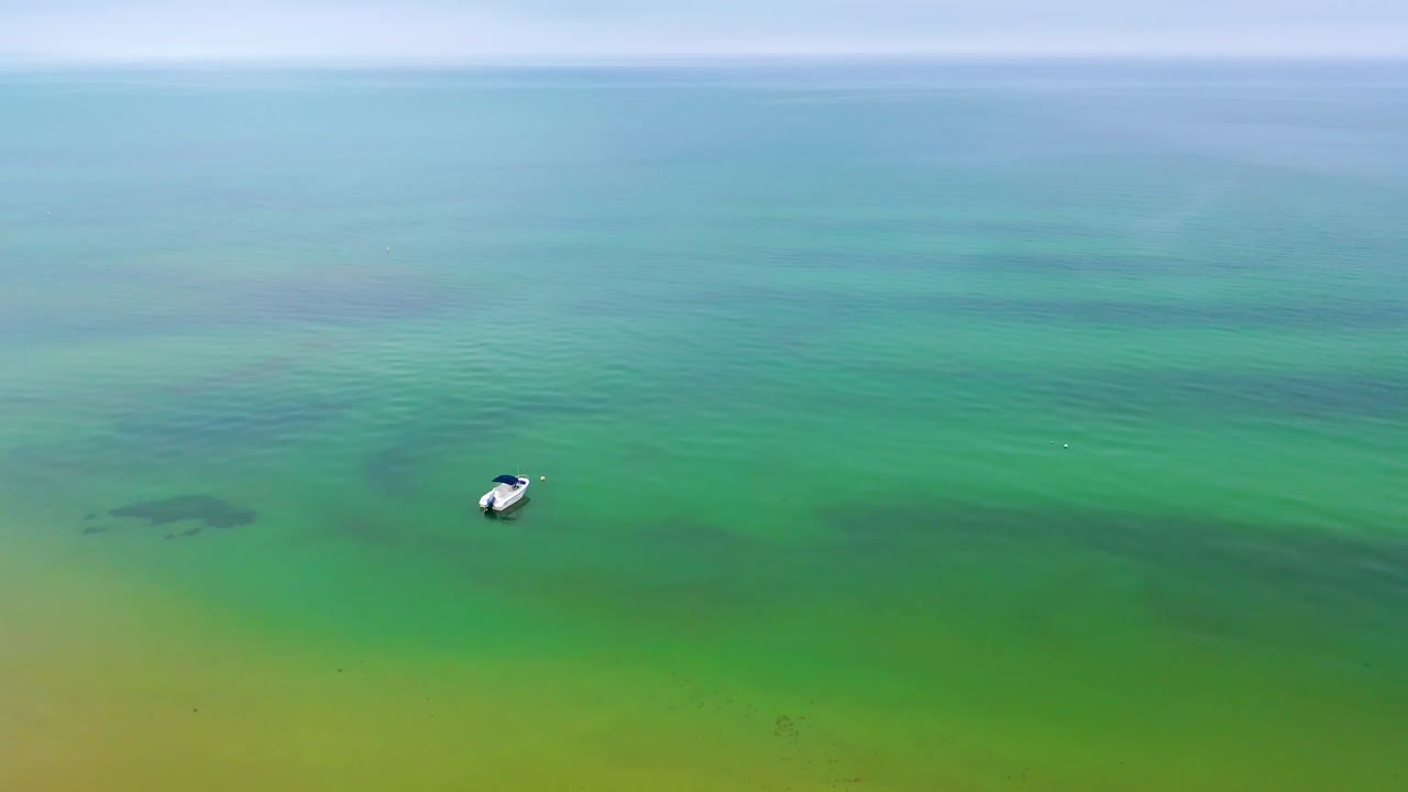 High-altitude drone footage of a boat floating on calm green-blue ocean waters, with soft ripples stretching across the surface under a cloudy horizon in the distance