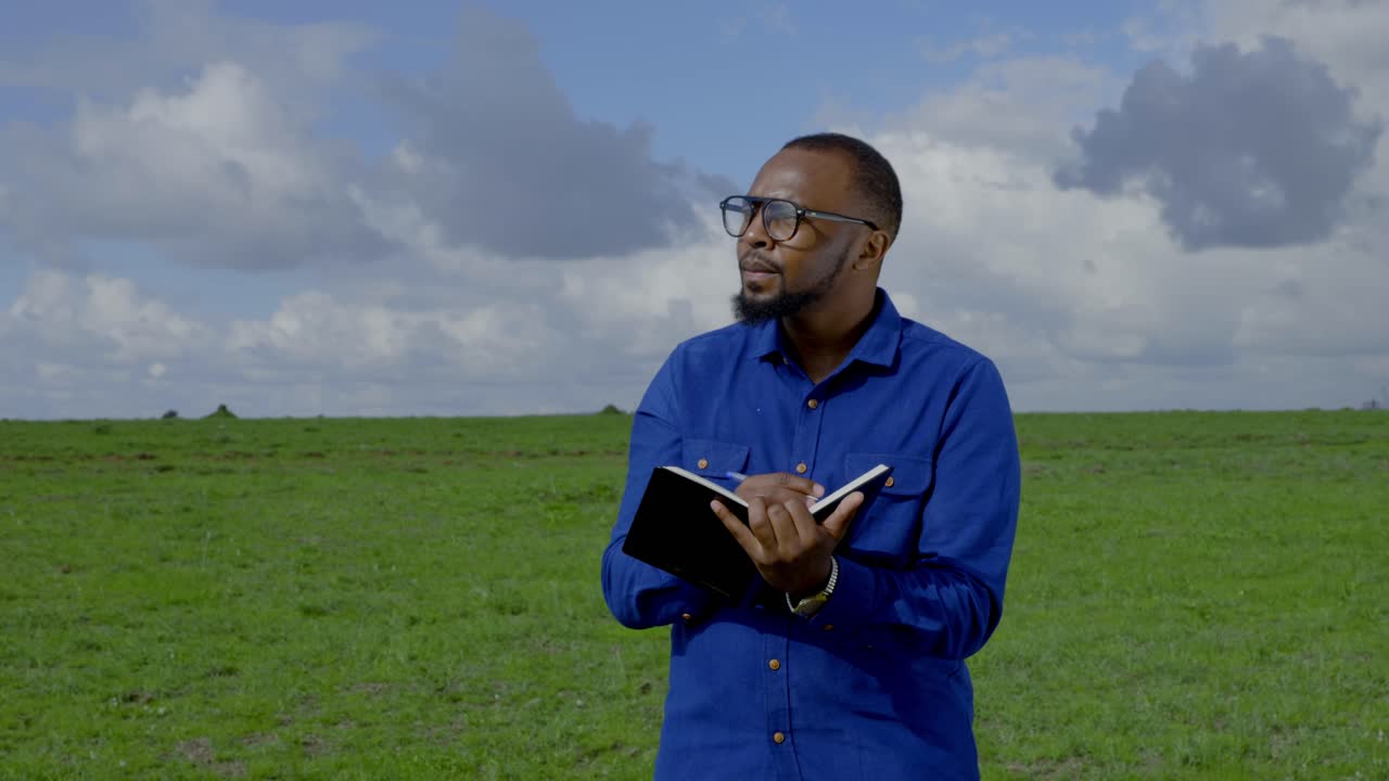 A black man stands in a meadow counting something and taking notes in a book
