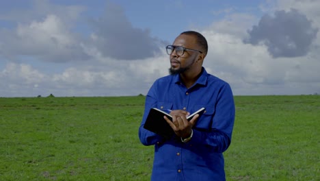 a black man stands in a meadow counting something and taking notes in a book