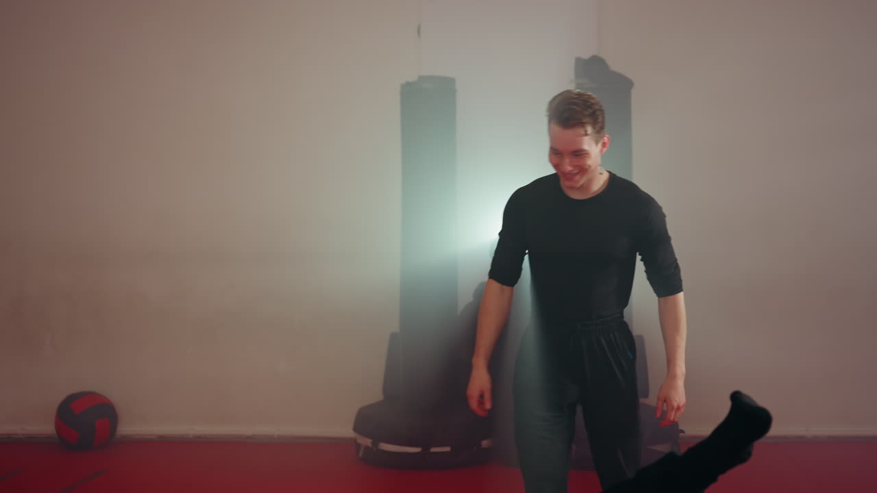 Two men training martial arts in gym, one man falling while holding shield during sparring, other standing ready, red mats on floor, foggy atmosphere, punching bags in background, bright spotlight glare