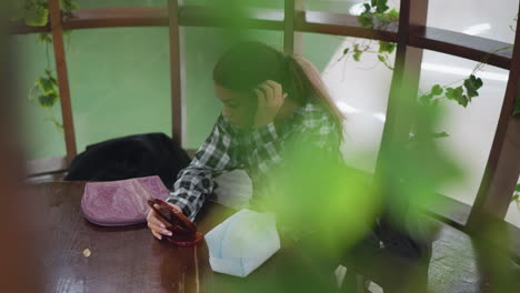 young girl adjusting her hair while looking into mirror, seated in a cozy, nature-inspired indoor space, green plants decorate the area, creating a peaceful ambiance as she focuses on her reflection