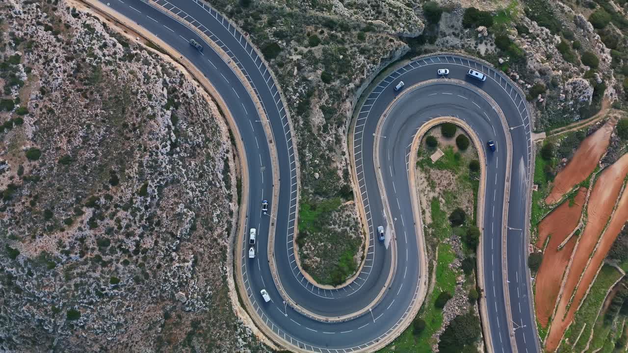 Vista aérea com inclinação descendente de uma estrada sinuosa em Mellieha, Malta, com veículos percorrendo curvas sinuosas através de uma paisagem mediterrânea rochosa e verde perto da costa.