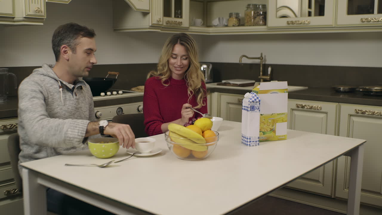 Una coppia seduta a un tavolo luminoso in cucina si gode la colazione. Ridono insieme mentre mangiano frutta e sorseggiano bevande calde in un'atmosfera rilassata.