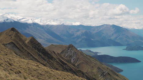 Golden-grass-on-spiky-mountains-with-deep-blue-lake-and-snowy-peaks-in-the-background