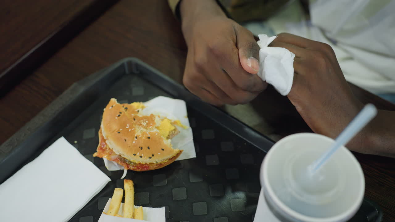 Overhead view of man cleaning fingers with napkin while seated at table with burger, fries, and drink cup, casual dining setting highlighting hygiene, lifestyle, and everyday eating detail indoors