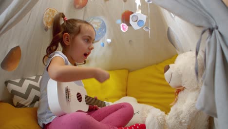 cute preschool girl teaches a teddy bear to play the guitar.