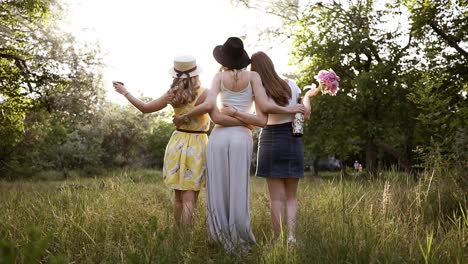 rare view of a group of three young nice girls embrace themselves looking at perspective. girl in the middle is holding a wine bottle. green meadow