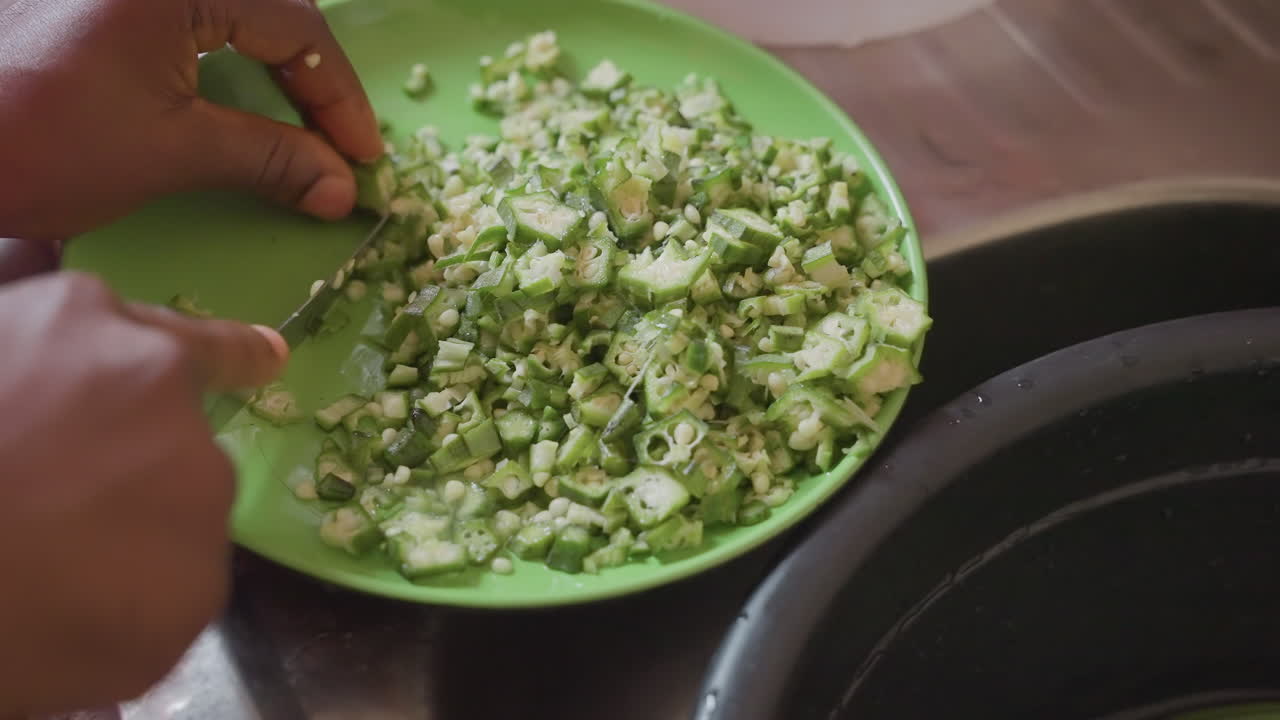 Mani nere che tagliano l'okra su un piatto verde, preparazione in cucina in primo piano da parte di un cuoco casalingo nero che usa un coltello per tritare l'okra in piccoli pezzi sopra il lavello, consistenza verde brillante, verdura fresca, ritmo delicato, famiglia