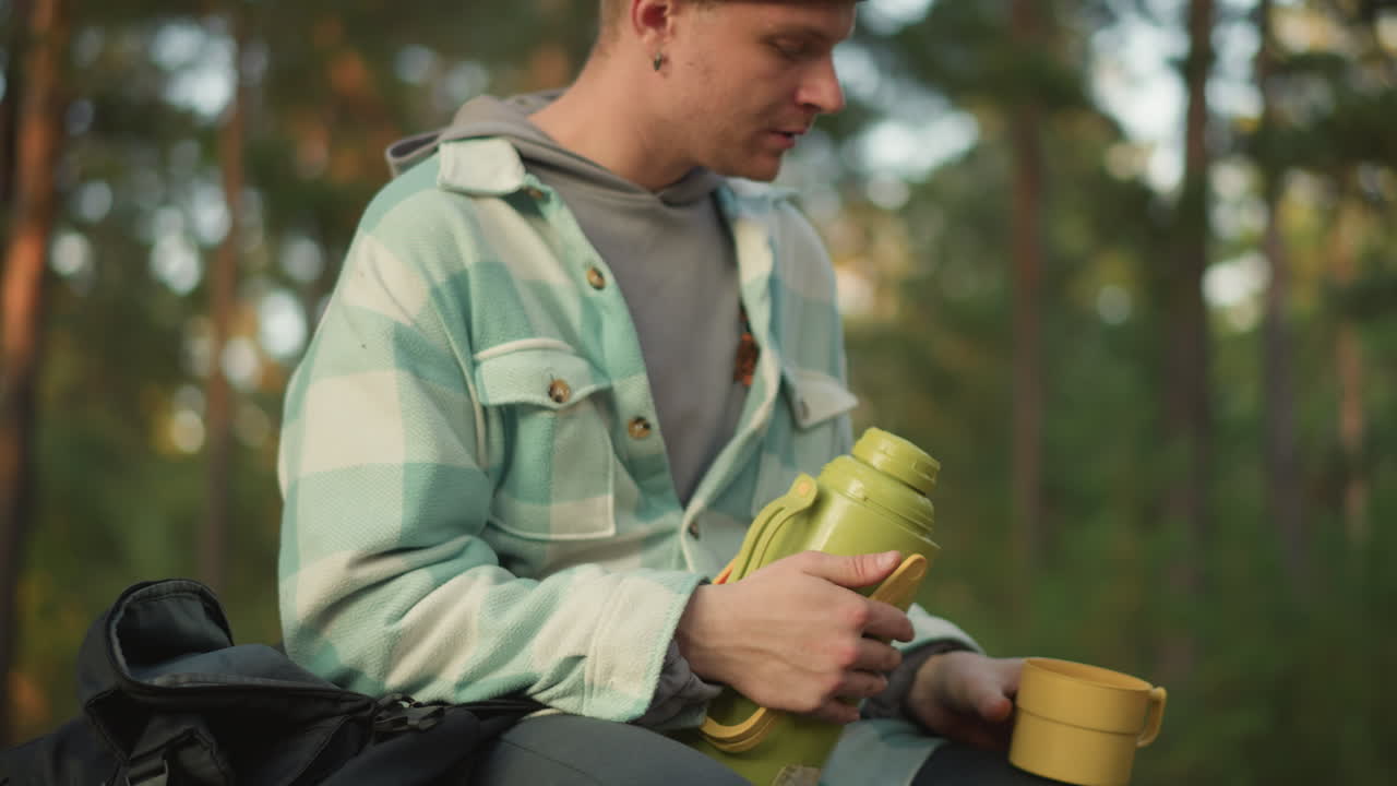 Viaggiatore si riposa serenamente, Sereno zaino in spalla sorseggia dal thermos in mezzo a un tranquillo scenario naturale, Viaggiatore rilassato testa attentamente il coperchio del thermos prima di godersi una bevanda calda sotto i pini illuminati dal sole al tramonto