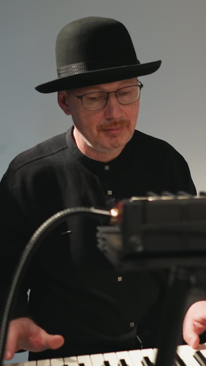 검은 모자, 안경, 검은 옷을 입은 키보디스트가 앉아서 회색 배경에 빨간 플러 키보드를 연주하며 자신의 공연에 집중합니다.