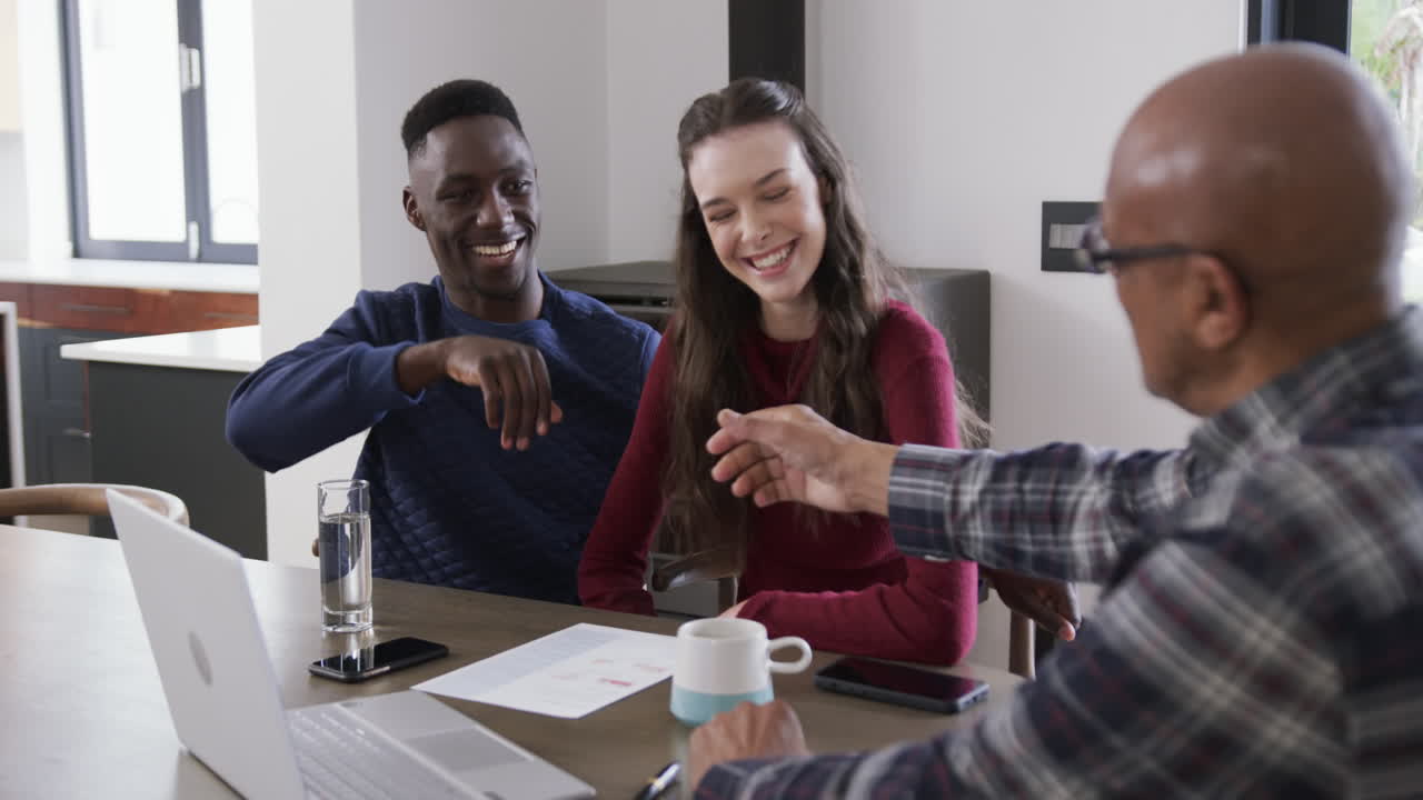 Happy diverse couple and male financial advisor using laptop shaking hands at home,slow motion