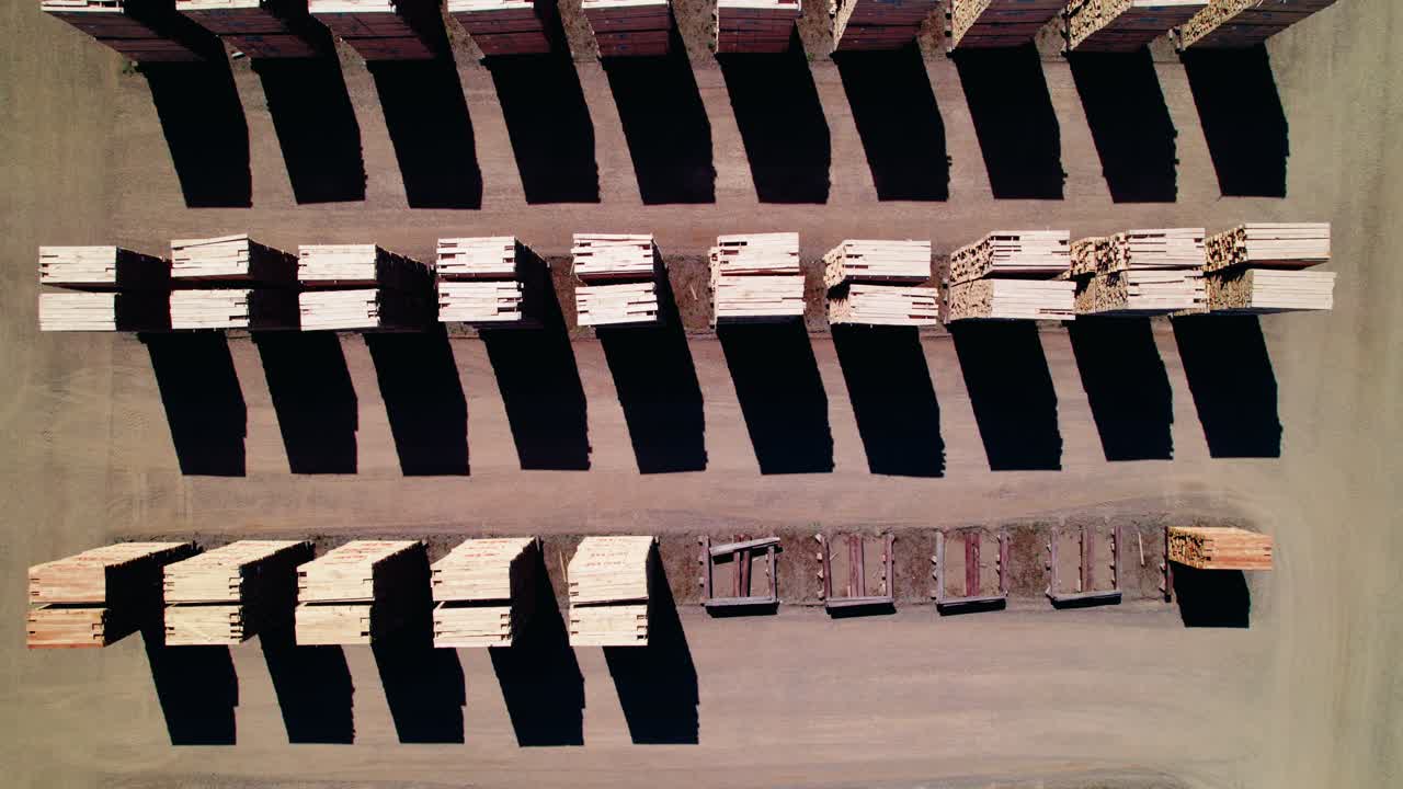 Top-down drone footage captures geometric timber harvest stacks casting crisp shadows across an open yard at a milling facility in Iron River, Michigan.