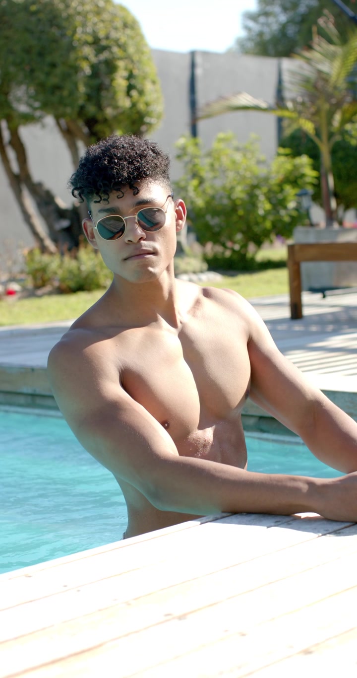 Video verticale: Uomo afroamericano a torso nudo con occhiali da sole che sente caldo a bordo piscina, sorseggiando da un bicchiere.