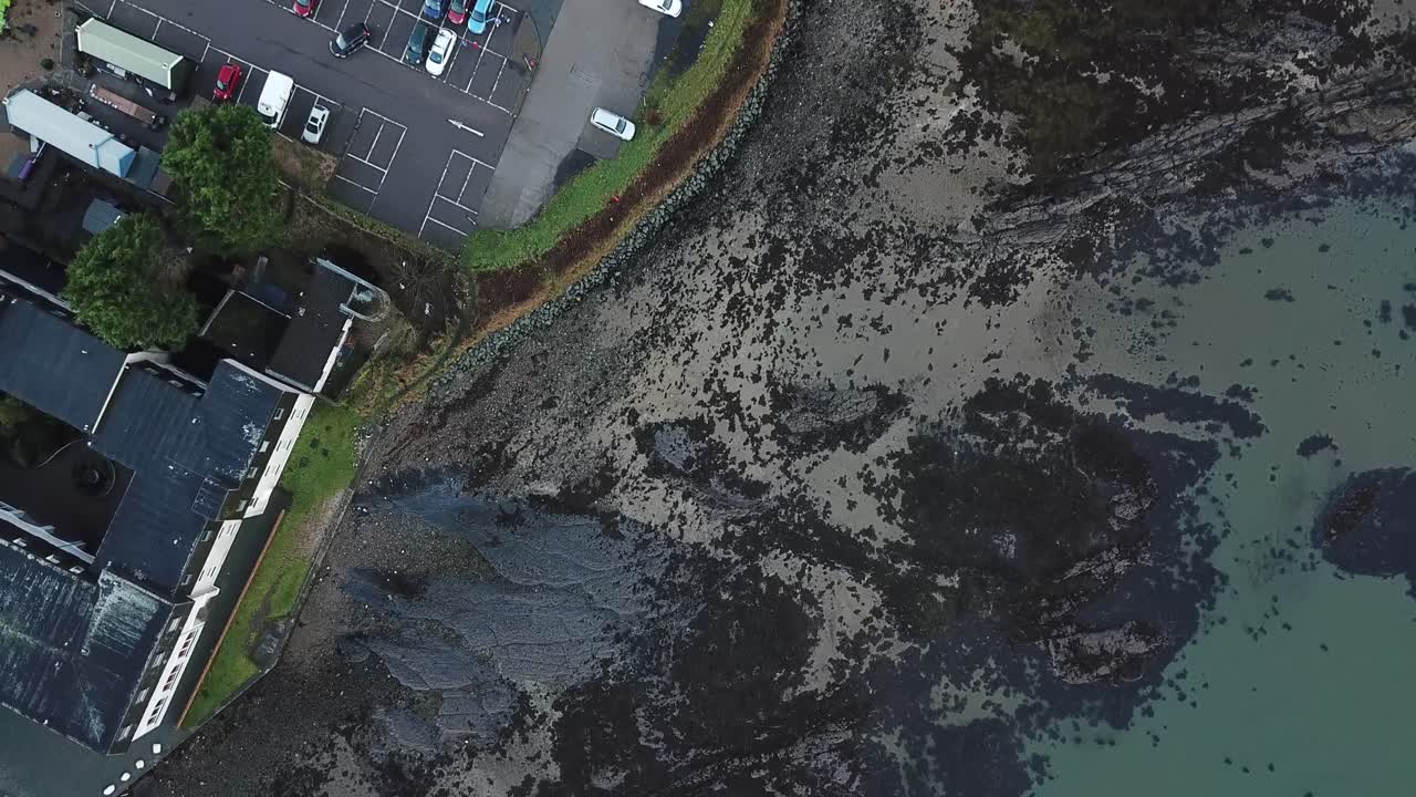 Aerial birds eye flyover of a beach with turquoise water, a car park and then a small village