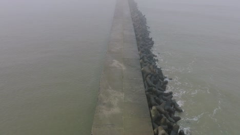 aerial establishing view of port of liepaja concrete pier, baltic sea coastline , foggy day with dense mist, moody feeling, big storm waves splashing, birdseye drone shot moving back, tilt up