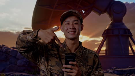 close up of asian man soldier smiling and pointing to smartphone in his hand while standing with satellite dish