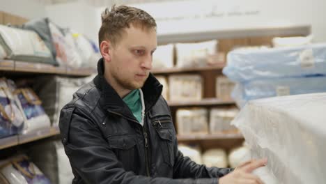 young man is choosing a mattress in a big furniture shop or supermarket. he is checking its elasticity, memory foam by pressing on it