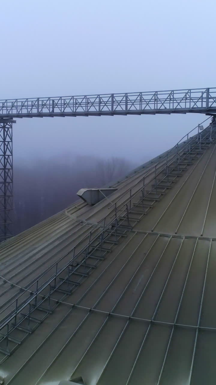 Dusty top of a huge silo tank at modern elevator. Drone shot slow motion over the silver granary tank on winter foggy weather. Vertical video
