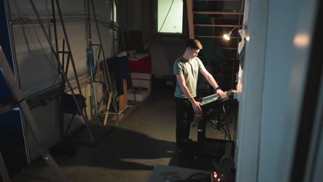 man in casual clothes stands in dim industrial workspace operating touchscreen control panel under warm desk lamp beside bulletin board filled with notes, while cables and ladders surround the scene