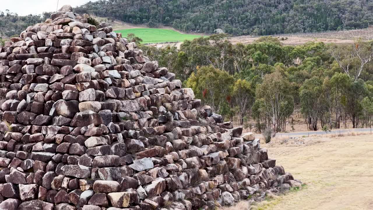 Camera pans across a large stone pyramid structure in a dry, grassy field with forested hills in the background under natural daylight