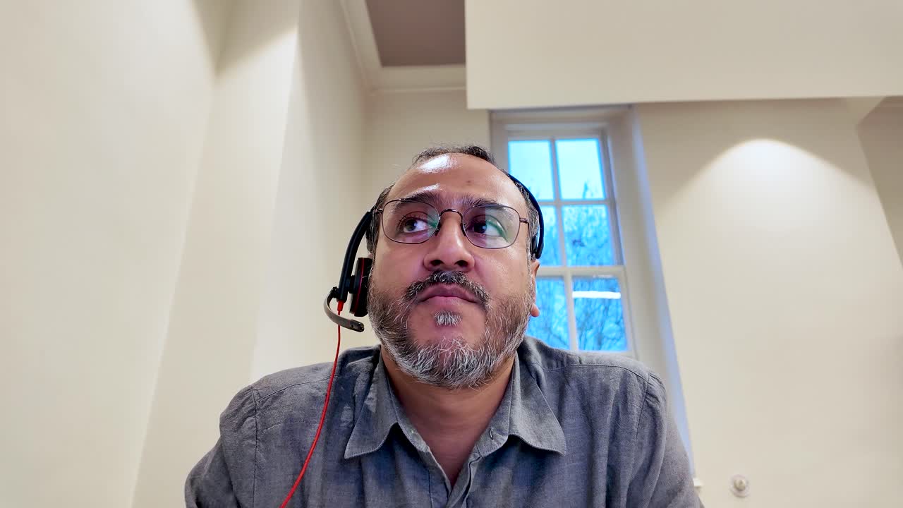Sørasiatisk mann med headset på kontor som telependler og jobber eksternt, konfererer og deltar på et virtuelt møte innendørs
