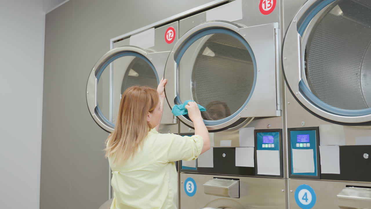 Laundry worker in yellow shirt seen from behind wiping surface door of industrial washing machine with soapy towel, reflection visible on glass, maintaining cleanliness in laundromat environment