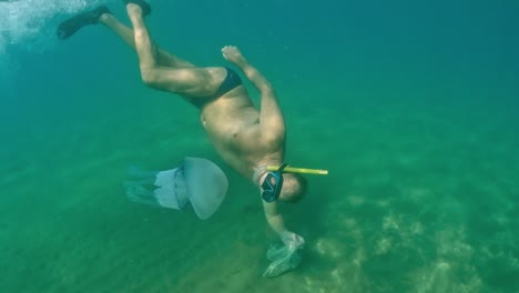 Vista-Subacuática-De-Un-Hombre-Adulto-Buceando-Para-Ver-La-Medusa-Medusa-Rhizostoma-Pulmo-Mientras-Sostiene-Una-Bolsa-Para-La-Pesca-De-Almejas-Y-Mejillones