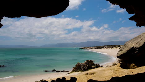 Pristine-stretch-of-white-secluded-beach-with-alluring-blue-water,-beautiful-sky-on-sunny-day,-panning-shot-from-cave