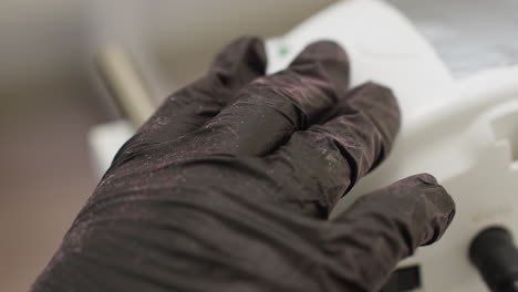 close-up view of gloved hand adjusting dial on white electric tool, covered in fine pink dust particles, indicating active use in cosmetic or nail care procedure in clinical workspace setting
