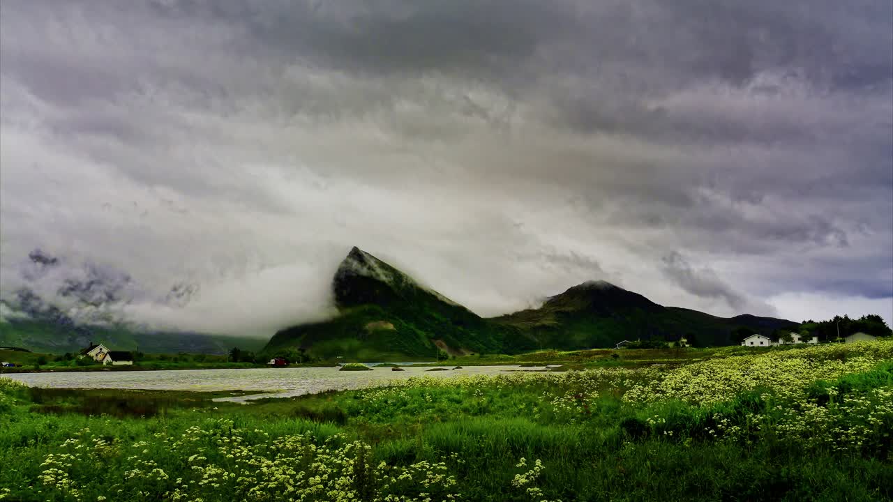 Approaching storm over the village by the mountains and lake surrounded by lush meadow. Time Lapse. Lofoten Islands, Northern Norway