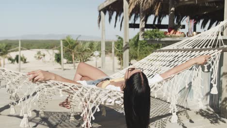 Entspannte-Hispanische-Frau-Mit-Sonnenbrille,-Die-In-Der-Hängematte-Am-Sonnigen-Strand-Liegt,-Zeitlupe