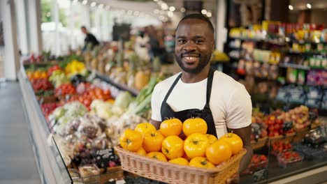portrait of a black-skinned man in a brown t-shirt and black apron posing with a box of yellow tomatoes in his hands in a supermarket