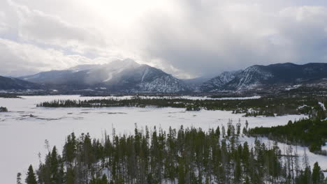 aerial moving left and up to reveal icy, frozen lake surrounded by snow and green pine trees with large snowy ski town mountains in the background near silverthorne and frisco colorado