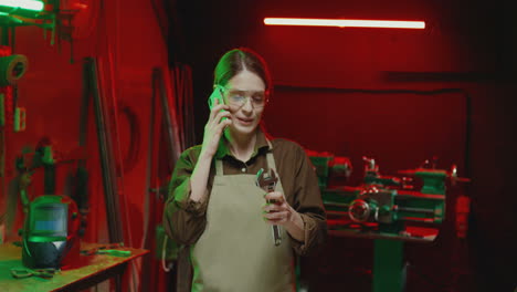 Cheerful-female-welder-in-apron-and-goggles-talking-on-phone-at-work-in-workshop