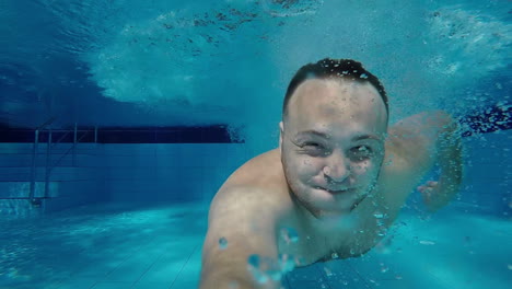 a man swims under the water in the pool with his eyes open looking into the camera underwater selfie