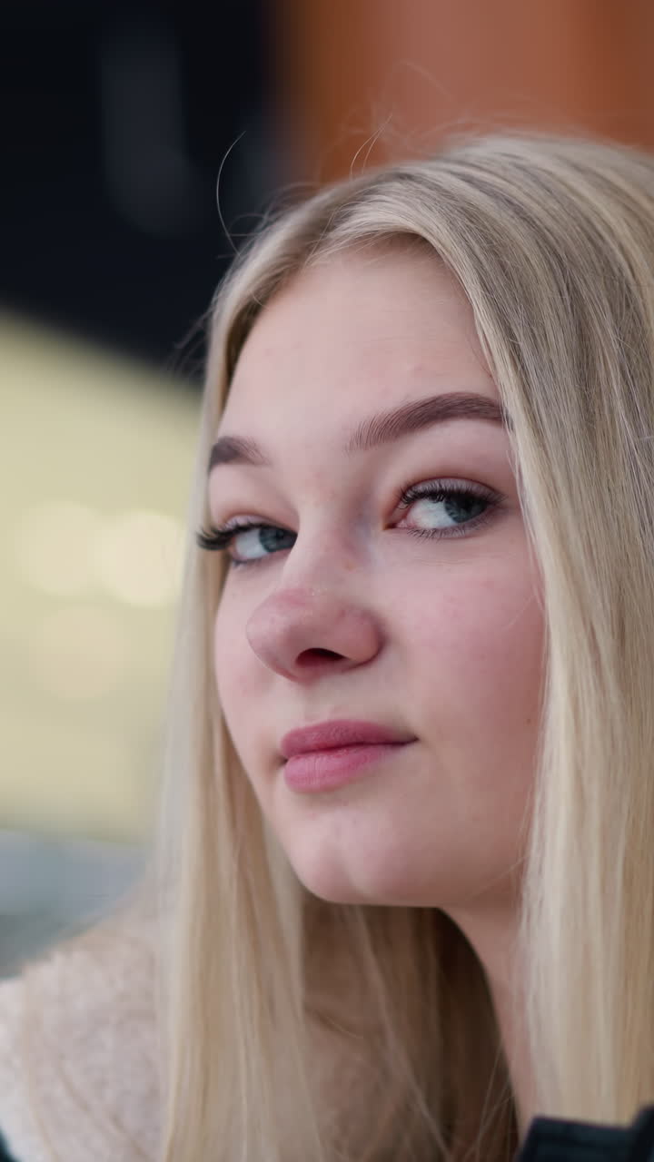 dama elegante con cabello rubio largo y sonrisa cálida mirando a su alrededor en el centro comercial, ella está usando una elegante chaqueta de invierno, de pie cerca de la barandilla de vidrio con fondo borroso