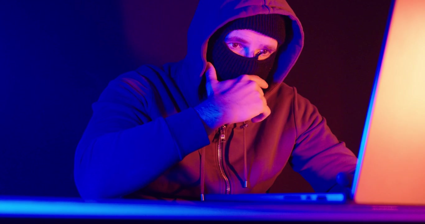 Persona mascherata con una felpa con cappuccio che usa un laptop in un ambiente buio con un'illuminazione al neon blu e rossa drammatica. Concetto di cybersecurity, cybercrime, hacking, minacce digitali e sicurezza online.