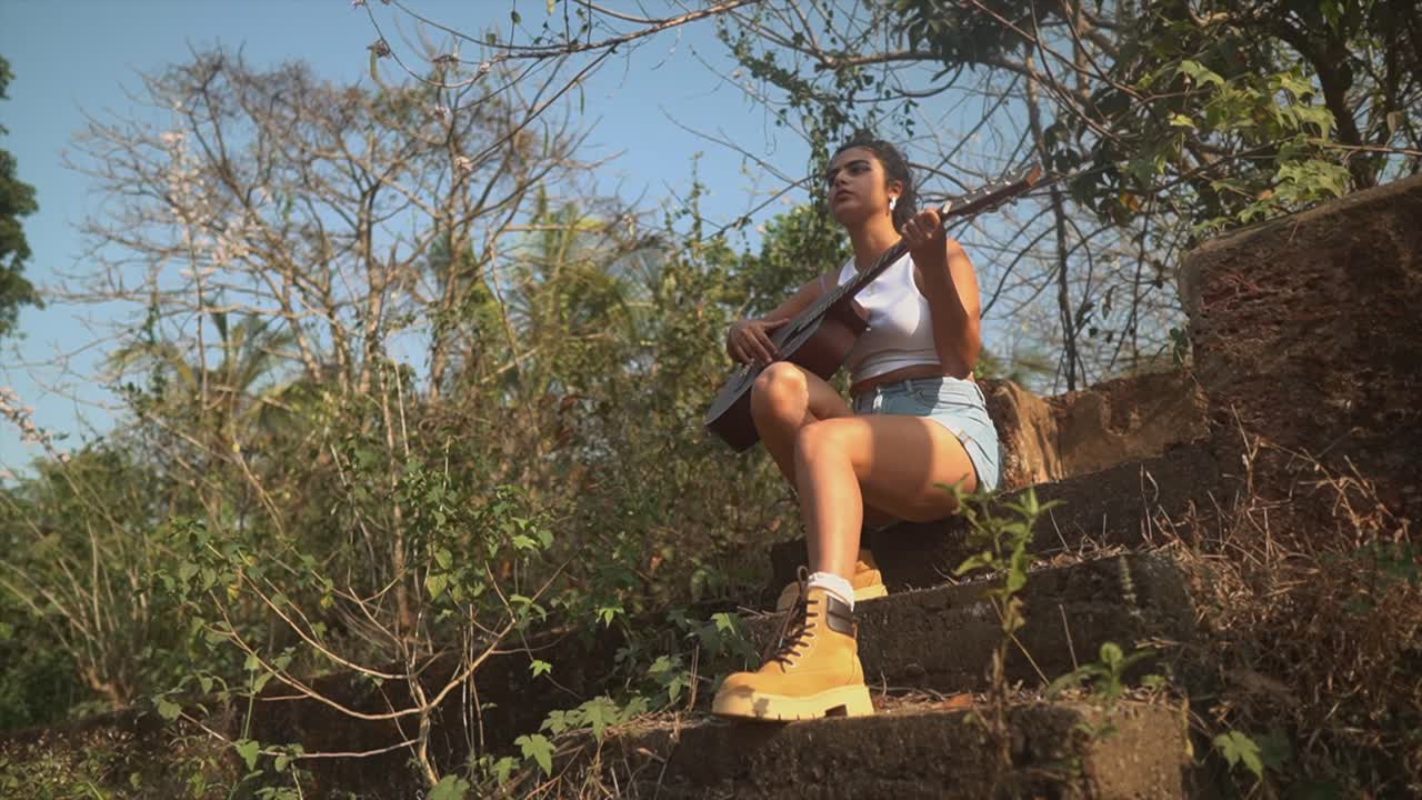 Inquadratura dal basso di una bella ragazza che suona la chitarra sotto un albero, illuminata dal sole e circondata da alberi e arbusti, seduta su delle scale.